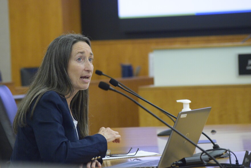 A woman with brunette hair sits at a desk with microphones around her. She is wearing a blue blazer and white shirt.