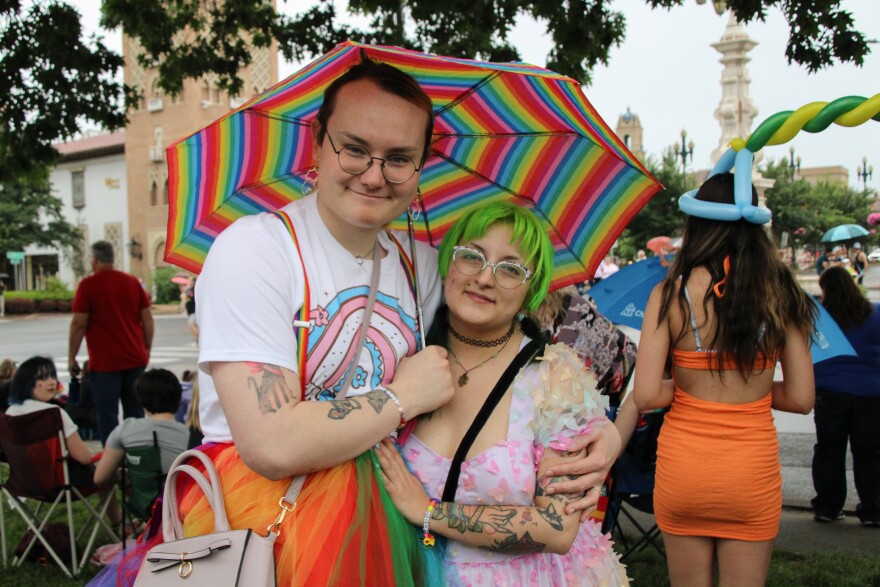 A woman in a rainbow tutu holds a rainbow umbrella and has her arm around another woman with green hair who is wearing a rainbow dress with butterflies on it.