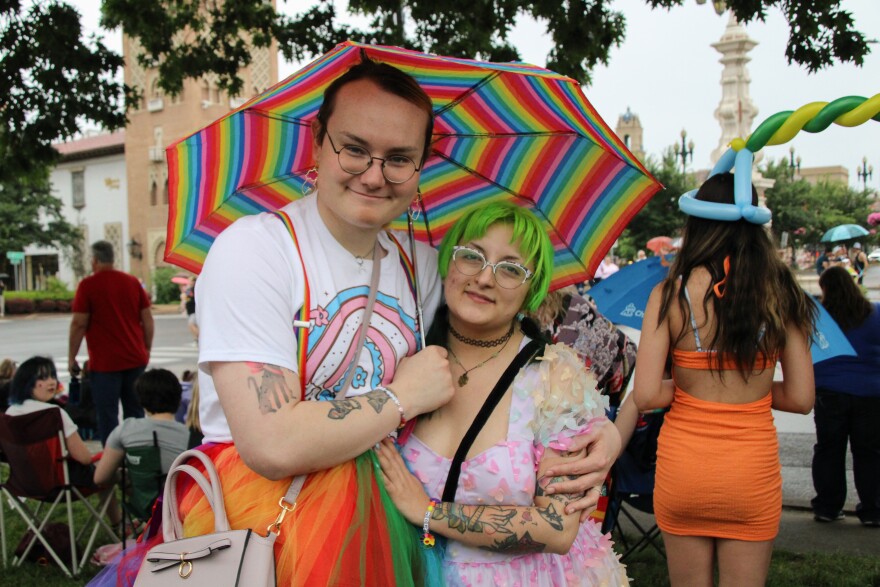  A woman in a rainbow tutu holds a rainbow umbrella and has her arm around another woman with green hair who is wearing a rainbow dress with butterflies on it.