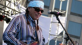 A man in a blue bandana and sunglasses plays the electric guitar in front of a mic stand