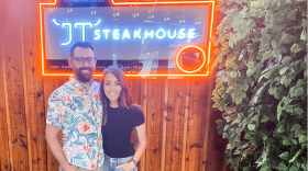 Terrill Trask, a man in black-rimmed glasses, light beige jeans and a floral shirt with a beard, and Jess Trask, a woman with brown hair in a black t-shirt and ripped blue jeans, smile with their arms around one another in front of a sign that reads “JT Steakhouse.”