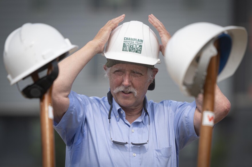 A man puts on a white hard hat. In the foreground are shovel handles with hard hats balanced on top
