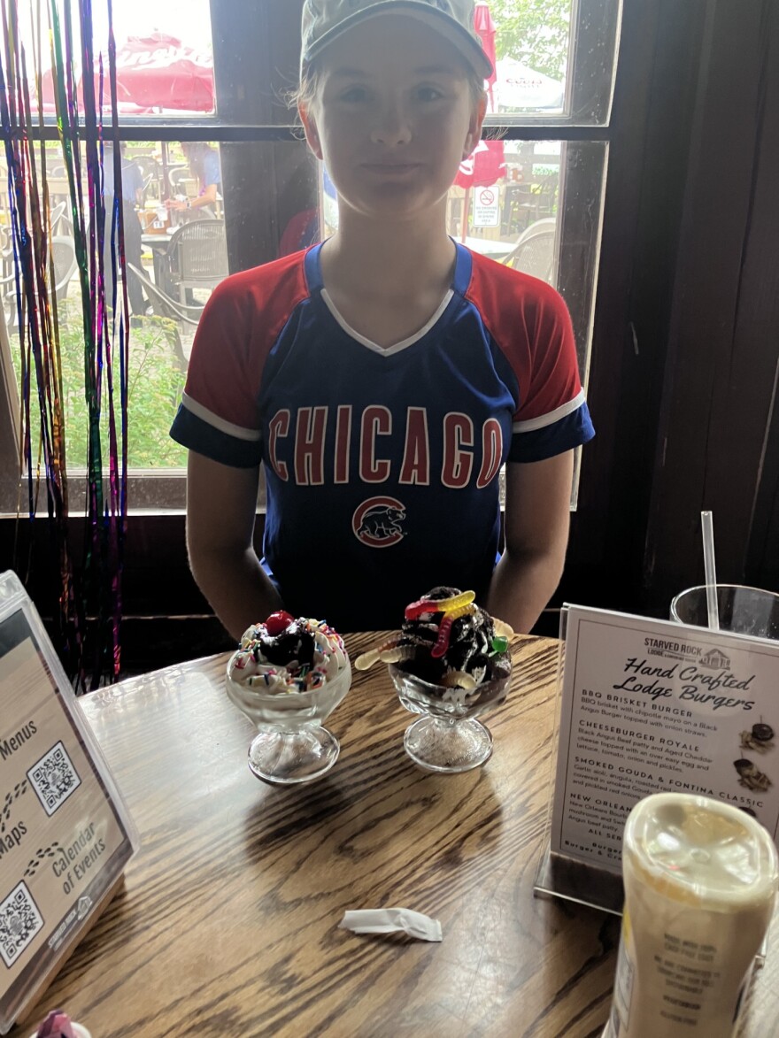 A girl sits at a table and in front of a window, there are two dishes of ice cream on the table. 