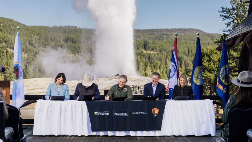 Five people sit at a long table signing agreements. The table has cover with the logos for Yellowstone Forever, Blackstone, Tallgrass, and the National Park Service. There are also state and federal flags behind them. In the background, you can see a geyser erupting in the heavily forested area.