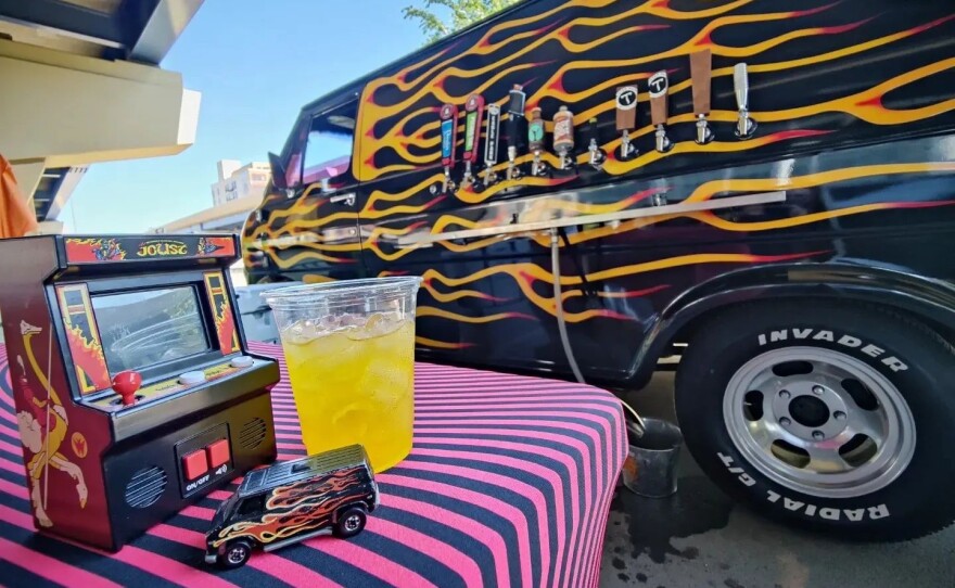A vintage van with flames and tap lines on the side, and a table in the foreground with a miniature arcade cabinet, drink and Hot Wheels toy van.