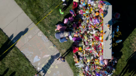 An aerial view of a memorial of flowers, balloons, and sidewalk chalk near a grassy area.