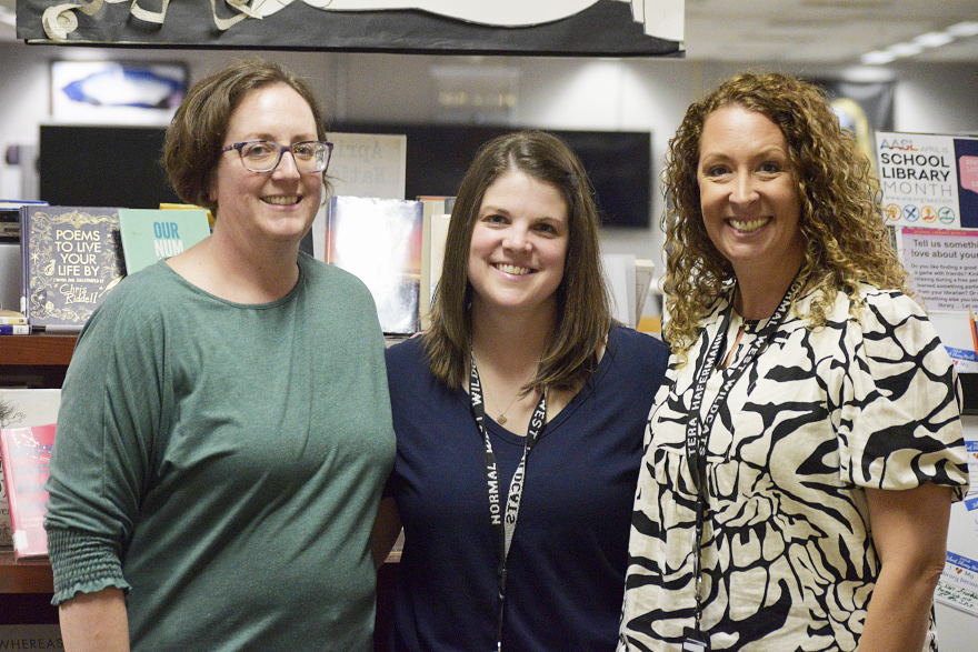 Three women smile in front of a bookshelf in a library.