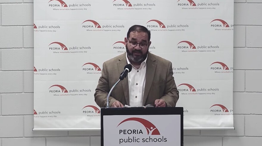 A man in a tan blazer speaks at a podium with a microphone. The podium and backdrop display the Peoria Public Schools logo and name repeatedly.