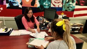 A woman sits behind a desk looking through papers as a young woman sits across from her filling out paperwork on a clipboard.