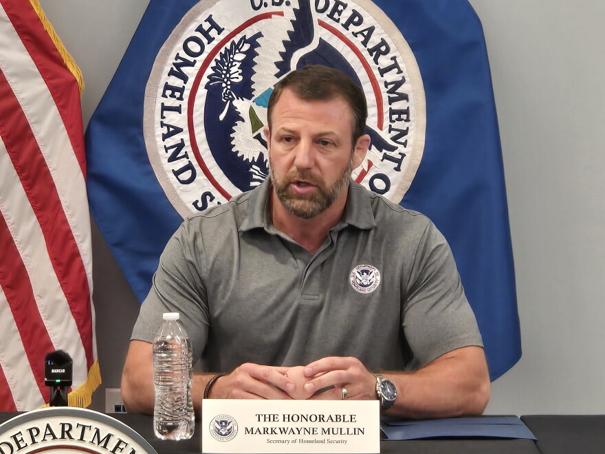 man in grey polo shirt sits at a table with flags behind him