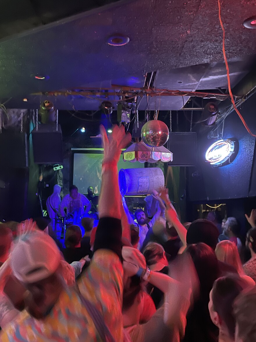 A view of a band on a stage taken from behind the audience with many audience members raising their hands in the air