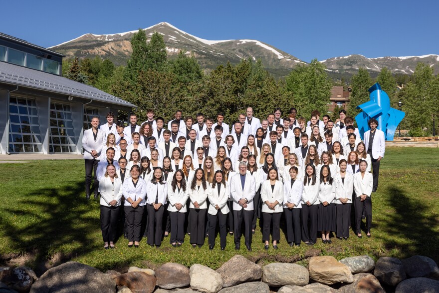 About 80 college-age musicians who are part of the National Repertory Orchestra in Breckenridge, stand in a group outside. They all wear black slacks, dark shirts, and white jackets. There are mountains and a blue sculpture behind the group.