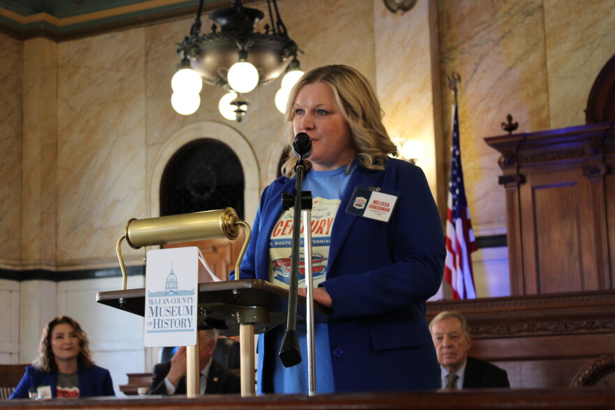 A woman in a blue blazer speaks at a podium labeled “McLean County Museum of History” in a historic courtroom, with an American flag and others seated behind her.