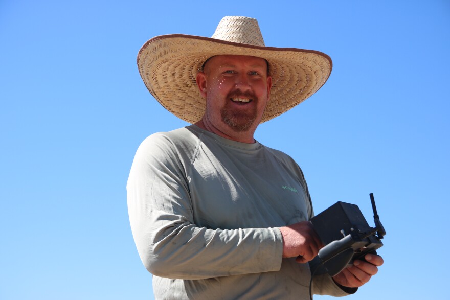 A man with a beard in a wide-brimmed, wicker sunhat and a grey, long-sleeved shirt looks and smiles. He is holding a square drone remote in his hands.