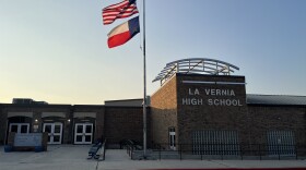 A school building with benches, doors, a sign that reads "La Vernia High School" and a flag pole flying the U.S. and Texas flags.