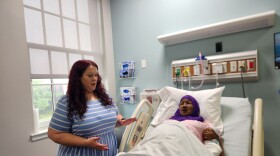 A woman in a striped blue dress stands next to a hospital bed. A hijab-wearing mannequin designed to simulate birth lies in the hospital bed with a mannequin designed to look like a newborn child.