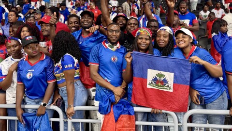 A large crowd in a stadium wearing blue and red jerseys, with two people holding a Haitian flag. 