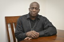 A man sits at a wooden table. He is wearing all black with a silver chain around his neck. His hands are folded before him and he is looking at the camera.