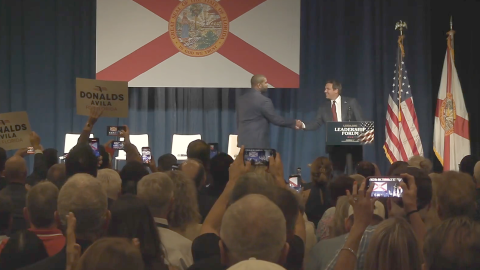 Florida Gov. Ron DeSantis shakes hands with U.S. Rep. Byron Donalds onstage in front of a large Florida flag as audience members watch and take photos.