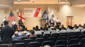 Coast guard graduates sit in the front two rows as an office speaks from a podium on the stage. 
