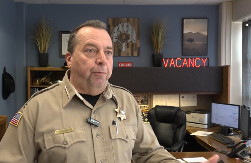 A man wearing a tan uniform and star-shaped sheriff badge speaks in his office. It's decorated with a neon sign that says "Vacancy" and some artwork of bison and a tree.