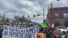 Protesters walk around an intersection. In fron of them, two people hold a white banner with blue letters that reads "NO DEPORTATIONS! NO DEPORTACIONES!" 