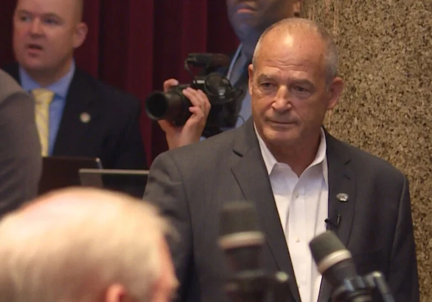 Gary Pinkel is seen in front of microphones. There is a photographer in the background with a man in a suit. Pinkel has a neutral expression.  