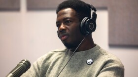 Kwabi Amoah-Forson sits in front of a microphone. He wears a pair of headphones and peace sign pin. He appears to be listening intently and with curiosity.