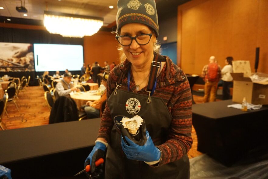 Margo Reveil of Jakolof Bay Oyster Company holds a shucked oyster at a March 10, 2026 reception at the Mariculture Conference of Alaska in Anchorage. Oysters in Alaska grow more slowly than those in farther-south locations, but the final product is premium, an expert from New York told the conference audience.