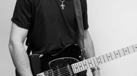 Black-and-white portrait of a young man wearing sunglasses, a T-shirt, and a cross necklace while holding a black electric guitar against a plain background.