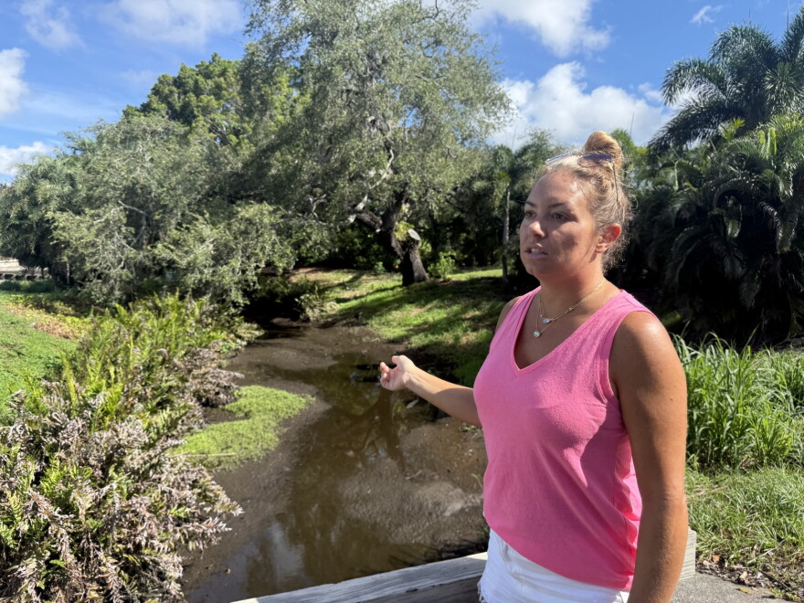Woman in pink shirt stands outside pointing at a creek 