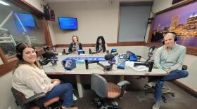 Four people wearing headphones sit at a table in a radio talk studio: a woman front left has long brown hair and is wearing a beige shirt, blue jeans and brown boots; a man front right has short dark hair and is wearing a light green button-down shirt, blue jeans and sneakers; a woman back left has long blonde hair and is wearing a black zip-up fleece over a grey t-shirt; a woman back right has long black and green dreadlocks and is wearing glasses and a pink shirt.