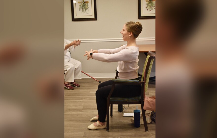 A woman wearing a pale pink sweater, leggings and ballet slippers sits in a chair in front of a class, leading them ballet arm positions.
