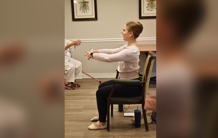 A woman wearing a pale pink sweater, leggings and ballet slippers sits in a chair in front of a class, leading them ballet arm positions. 