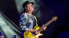 Carlos Santana dressed in white and blue playing guitar on blue background