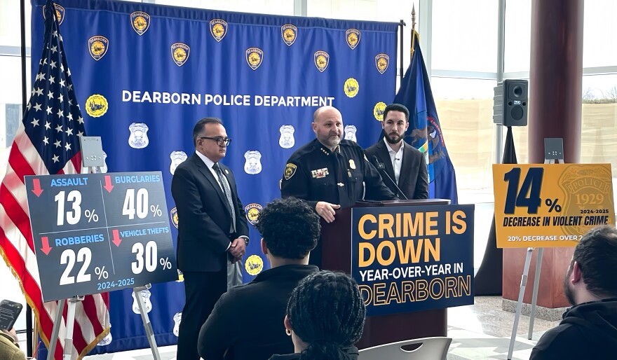Three men, two in dark suits and one in a police uniform, stand in front of a podium with a sign that reads "CRIME IS DOWN year over year in Dearborn." Two other signs with percentages are on either side of the group, along with an American and Michigan flag. 