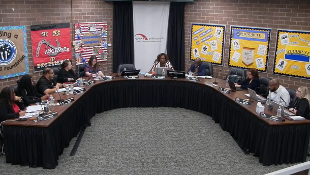A group of people sit around a large U-shaped table in a meeting room with bulletin boards on the walls and “Peoria Public Schools” on a banner behind the center person.