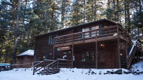 The Fort Abercrombie ranger station under a light blanket of snow in March 2026. It is a two story building with dark brown siding and green accents on the windows. Behind the station, sun filters through spruce trees.