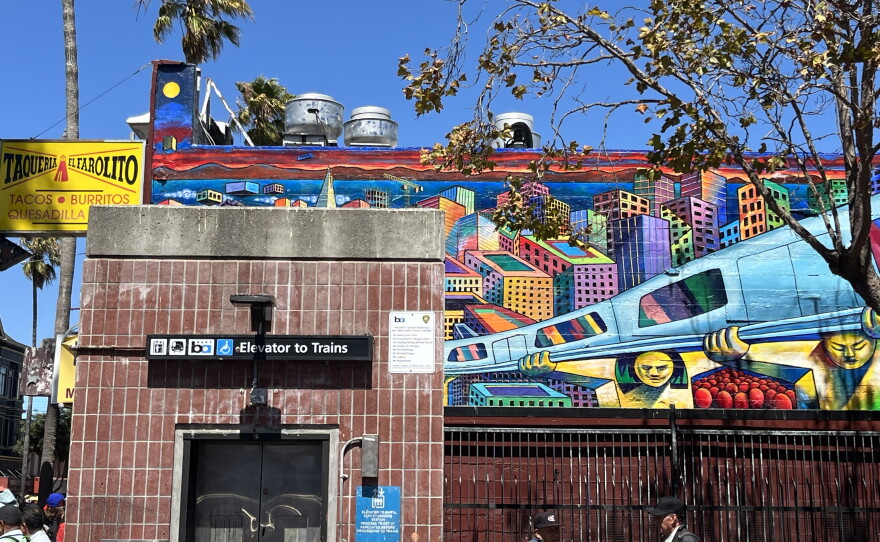 A mural on the wall of El Farolito on Mission Street, facing the northeast corner of the 24th Street BART plaza, by artists Michael J. Rios, Richard Montes, and Tony Machado. Depicts golden figures holding up a silver BART train against the colorful backdrop of the city. 