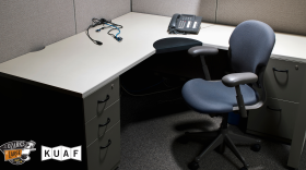 An empty office cubicle with a gray desk, a black office chair, a telephone, and tangled cords.