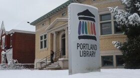 A snow-covered sign that says "Portland Library" at the front entrance of the snowy building
