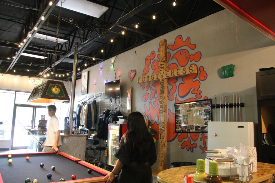 Two kids play pool at a pool table. Behind them is a wooden cross that says "Forgiveness" across one of the beams. On the wall is some pink graffiti and lights. There's a welcome desk behind them and the front entrance doors. To the right is a bar with food.