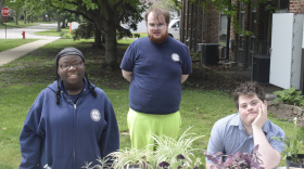 Three students smile behind a row of plants.