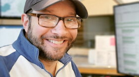 Mitch Teich, pictured in a quarter-zip, tortoise shell glasses and a baseball cap smiles in front of radio equipment. 