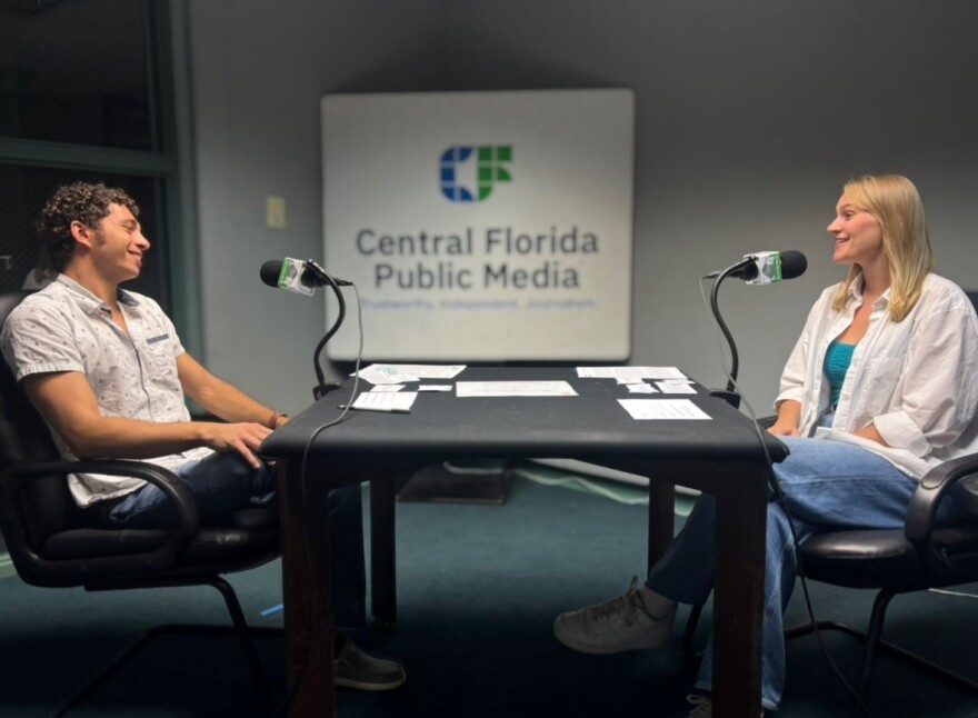 A man sits on the left looking to the woman sitting on the right. They both have microphones in front of them