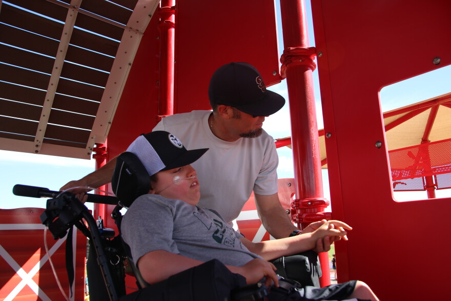 A boy in a wheelchair wearing a baseball cap and a gray tee looks out the window of a red structure. An older man wearing a black baseball cap and a white tee leans over him and looks out the window with him.