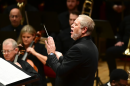 Man in suit with grey hair conducts group of people holding instruments 