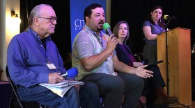  A group of people sit on a stage, one person speaks into a microphone, one person is at a podium.