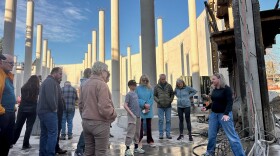 A woman speaks to a group of people standing inside a massive, roofless round structure filled with columns. 