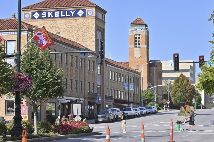 Exterior photo of a city street shows two people crossing the street. Buildings can be seen in the background with cars parallel parked in front of them.
